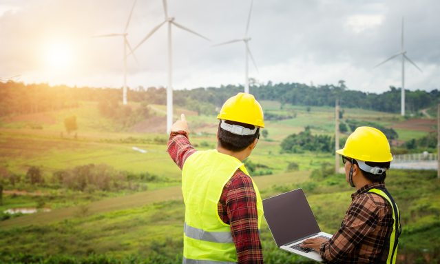 Two people in hard hards and personal protective gear pointing at wind turbines