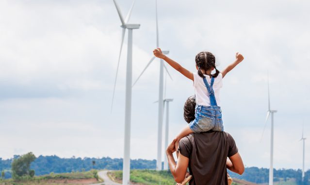 A man with a child on his shoulders, looking at wind tourbines